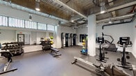 Located at 11 E Pleasant St in Amherst, Massachusetts, this fitness center offers a spacious, single-floor layout designed to accommodate a comprehensive range of workout activities. The space features an industrial aesthetic with exposed ductwork, structural columns, and open ceilings fitted with track and pendant lighting, creating a functional yet modern atmosphere.

The fitness floor is organized into distinct training zones. A dedicated cardio area houses multiple treadmills and elliptical machines, providing ample space for aerobic exercise. A strength training section features dual cable resistance machines positioned alongside a large wall-mounted mirror, allowing users to monitor their form during workouts. A free weights zone includes a full dumbbell rack and adjustable benches, while medicine balls, slam balls, and resistance bands are neatly organized on dedicated storage racks.

Additional cardio equipment, including a stationary bike and a rowing machine, occupies a separate area near the glass-paneled entry doors, which allow natural light to filter into the space. Clerestory windows along one wall further brighten the interior, contributing to an open and energizing environment. The durable rubber flooring throughout provides safety and comfort underfoot. The gym's layout ensures clear, unobstructed movement between zones, making it practical and accessible for a variety of fitness routines.