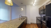 Situated at 8 E Long St in the heart of downtown Columbus, Ohio, this single-floor residence offers a well-designed layout with contemporary finishes throughout. The open-concept kitchen serves as a central gathering point, featuring a granite-topped island with a double undermount sink, dark espresso cabinetry, pendant lighting, and a track lighting system. Black appliances including a refrigerator, range, and microwave are neatly integrated into the cabinetry, complemented by recessed ceiling lighting that brightens the space.

Rich hardwood flooring extends through the main living areas, enhancing the warm, urban feel of the unit. Large, black-framed windows allow generous natural light to fill the living and bedroom spaces, offering views of the surrounding downtown cityscape. The bedroom area is spacious and transitions to an additional carpeted room, ideal for use as a second bedroom or home office.

The bathroom is finished with the same granite countertop seen in the kitchen, set into dark cabinetry with modern hardware, a framed mirror, and clean white tile flooring — creating a cohesive aesthetic. The overall design balances contemporary urban style with functional comfort, making it a practical and attractive residence in the heart of Columbus.