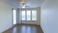 This single-floor space presents a clean, open layout with a welcoming and airy ambiance. The main living area is finished with rich hardwood-style flooring that runs throughout, adding warmth and a polished aesthetic to the space. Neutral-toned walls provide a versatile backdrop suited to a variety of interior styles.

Natural light is a defining feature of this space, with a set of three large windows fitted with horizontal blinds along one wall, complemented by an additional side window and what appears to be a glass-paneled door. Together, these openings draw in generous amounts of daylight and offer views of the surrounding greenery and outdoor environment.

A ceiling fan with an integrated light fixture is centrally mounted, contributing to both the functionality and comfort of the room by promoting air circulation and providing supplemental lighting. The clean white ceiling and smooth wall finishes give the interior a fresh, move-in-ready feel.

Overall, the space conveys a sense of openness and practicality, with thoughtful design elements that prioritize light, flow, and everyday comfort.