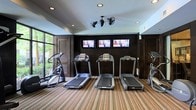 This fitness center is thoughtfully designed to provide a complete workout experience in a polished, comfortable setting. The space features a dark, sophisticated accent wall that serves as a bold backdrop for three wall-mounted flat-screen televisions, allowing guests to stay entertained during cardio sessions. A row of treadmills lines the center of the room, accompanied by an elliptical machine and additional strength or cardio equipment toward the perimeter. The striped, multi-toned carpeting adds a dynamic visual element underfoot while providing cushioning throughout the space. Overhead, recessed lighting is complemented by a distinctive dual-head ceiling fan, ensuring the room remains well-lit and comfortably ventilated. Large windows along one wall flood the space with natural light and offer views of the lush greenery outside, creating a refreshing atmosphere during workouts. Dark louvered doors on either side of the accent wall add architectural character and provide access to additional areas. The overall ambiance is energetic yet refined, striking a balance between function and design.