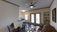 This single-floor space offers a well-appointed open layout that blends functional design with distinctive decorative touches. The living area is anchored by rich, dark hardwood flooring that extends throughout, lending warmth and a cohesive feel to the space. A multi-blade ceiling fan with an integrated light fixture is centrally mounted, providing both air circulation and ambient lighting.

Natural light enters through a set of French doors fitted with horizontal blinds, which appear to open onto an outdoor balcony or patio, and a secondary window nearby reinforces the bright, airy atmosphere. Crown molding along the upper walls adds a refined architectural detail.

A standout feature is the built-in wet bar or pass-through area, highlighted by a stone or granite countertop and a decorative wall treatment with an abstract, organic pattern — giving the space a creative, artistic character. Bar-height seating accompanies this area, making it ideal for casual entertaining.

Built-in shelving is visible on one wall, offering display and storage opportunities. The living area accommodates a seating arrangement centered around a glass-top coffee table, completing this inviting, versatile space.