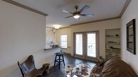 This single-floor space offers a well-appointed open layout that blends functional design with distinctive decorative touches. The living area is anchored by rich, dark hardwood flooring that extends throughout, lending warmth and a cohesive feel to the space. A multi-blade ceiling fan with an integrated light fixture is centrally mounted, providing both air circulation and ambient lighting.

Natural light enters through a set of French doors fitted with horizontal blinds, which appear to open onto an outdoor balcony or patio, and a secondary window nearby reinforces the bright, airy atmosphere. Crown molding along the upper walls adds a refined architectural detail.

A standout feature is the built-in wet bar or pass-through area, highlighted by a stone or granite countertop and a decorative wall treatment with an abstract, organic pattern — giving the space a creative, artistic character. Bar-height seating accompanies this area, making it ideal for casual entertaining.

Built-in shelving is visible on one wall, offering display and storage opportunities. The living area accommodates a seating arrangement centered around a glass-top coffee table, completing this inviting, versatile space.