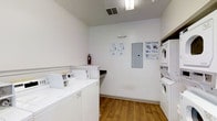 This shared laundry facility is designed with practicality and efficiency in mind. The room features a bright, clean aesthetic with crisp white walls and warm hardwood-style flooring that creates an inviting and hygienic environment. Multiple top-loading washing machines are neatly arranged along one side of the room, while a set of stacked dryer units lines the opposite wall, maximizing the use of available space. A built-in countertop surface provides a convenient area for sorting and folding laundry. The room is well-lit with overhead fluorescent lighting and benefits from a ceiling ventilation system to maintain air quality. An electrical panel is centrally mounted on the wall for easy access and maintenance. Instructional signage, including drying guides and service information, is posted on the walls to assist users. A fire extinguisher is also present, underscoring the facility's commitment to safety. The overall layout is thoughtfully organized to allow comfortable movement throughout the space, making it a fully functional and user-friendly laundry facility suitable for shared residential or commercial use.