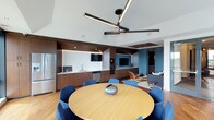 This single-floor space showcases a thoughtfully designed, open-plan layout that seamlessly integrates multiple functional zones. The main gathering area features warm hardwood flooring, a round wood dining table surrounded by blue upholstered chairs, and a modern ceiling fan with integrated linear lighting that anchors the space overhead. A tiered ceiling detail adds architectural depth.

The built-in kitchenette runs along one wall, finished in warm wood cabinetry with white countertops, a stainless steel sink, dishwasher, and microwave. A textured white tile backsplash and under-cabinet LED lighting add visual interest, while a wall-mounted TV is integrated seamlessly into the cabinetry. Open shelving provides decorative display opportunities. Adjacent to the kitchenette, a second media wall features a bold blue vertical tile accent and another mounted television, creating a dedicated lounge area with seating.

A distinct bar zone, separated by a transition in flooring to a teal herringbone tile, lines a wall of floor-to-ceiling black-framed windows. A long white high-top table with black stools fills this area, offering abundant natural light and elevated outdoor views. Contrasting yellow vertical accents punctuate the dark window framing, adding a vibrant design detail. Wall art and a sofa lounge area complete the space's versatile, contemporary character.