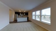 Located at 3381 Blue Heron Dr in Hilliard, Ohio, this single-floor residence offers an open-concept design that seamlessly connects the kitchen and living areas. The kitchen is appointed with dark cabinetry, stainless steel appliances including a refrigerator and range, and a central island with pendant lighting overhead, providing both functional workspace and a natural focal point. Dark hardwood-style flooring grounds the kitchen area, transitioning into plush neutral carpeting throughout the living space. Crown molding runs along the ceiling, lending a polished, finished look to the interior. The living area benefits from an abundance of natural light, courtesy of multiple large windows fitted with blinds, offering views of the surrounding outdoor greenery. The overall ambiance is clean, modern, and move-in ready, with a neutral color palette that provides a versatile backdrop for a variety of interior styles. The open layout promotes a sense of spaciousness, making the combined kitchen and living zone feel welcoming and functional for everyday living.