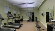 This single-level fitness room offers a practical and well-organized space designed to support a range of exercise routines. The walls are painted in a soft sage green, creating a calm and focused atmosphere conducive to working out. The room is illuminated by a combination of overhead fluorescent strip lighting and a ceiling fan, ensuring the space remains well-lit and ventilated during use.

The flooring features a neutral-toned, low-pile carpet that provides a comfortable surface throughout. On the cardio side, the room is equipped with multiple treadmills and at least one elliptical or recumbent cross-trainer, all from reputable fitness brands. For strength training, a multi-station cable and weight machine anchors one section of the room, accompanied by a flat workout bench and a set of free weights, offering versatility for both resistance and functional training.

Framed artwork adorns the walls, adding a refined touch to what is otherwise a utilitarian space. Horizontal blinds on the windows allow for light control and privacy. The overall layout is spacious enough to move freely between equipment, making this fitness room a well-rounded and accessible facility.