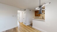 This thoughtfully designed 2-bedroom, 1-bathroom residence offers a bright and modern living experience across a single floor. Light wood flooring flows consistently throughout, creating a warm and cohesive aesthetic in every room.

The open kitchen-living area serves as the heart of the home, showcasing warm wood cabinetry, white quartz countertops, a tile backsplash, and a full suite of stainless steel appliances including a dishwasher and refrigerator. A peninsula with a built-in gas cooktop and a sleek stainless steel island range hood doubles as a casual dining or prep space, while track and pendant lighting add a modern finishing touch.

The spacious primary bedroom features light-toned wood flooring and a wide window fitted with horizontal blinds, filling the room with natural light. The second bedroom mirrors this brightness, complemented by a ceiling fan and an additional window with outdoor views.

The bathroom is cleanly appointed with a stone-look quartz vanity, warm wood cabinetry, matte black fixtures, and a marble-tiled shower surround. Two hallways connect the rooms fluidly, and a dedicated closet adds practical storage. White walls and clean lines throughout give this residence a fresh, move-in-ready appeal.