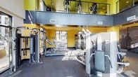 This two-story fitness center is designed to accommodate a full spectrum of workout needs within a vibrant, energetic environment. The lower level serves as the primary workout floor, featuring rubber flooring throughout and an extensive selection of strength-training machines, including multi-function cable systems, plate-loaded equipment, and dedicated benches. A row of treadmills equipped with individual media screens lines the perimeter, positioned alongside large windows that invite abundant natural light and provide views of the surrounding outdoor areas.

The space is visually defined by bold yellow walls and a teal ceiling accent, lending the facility a dynamic, modern character. Double-height ceilings soar above a portion of the main floor, creating an expansive, airy atmosphere. A staircase — decorated with a striking silhouette mural of athletic figures — connects the lower level to the open mezzanine above, where additional cardio equipment such as ellipticals and upright bikes are arranged along a railed overlook. The mezzanine is framed by a sleek metal cable railing system, offering an open sightline to the main floor below.

Large floor-to-ceiling windows on multiple walls flood both levels with natural light, further enhancing the sense of openness and scale throughout the facility.