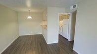 This single-floor residence offers a practical and well-organized layout designed to maximize functionality. The living and dining area is open and airy, featuring rich, dark hardwood-style flooring that runs throughout the main space, creating a warm and cohesive look. A textured popcorn ceiling adds a classic touch, while neutral beige walls provide a clean, versatile backdrop. A pendant light fixture hangs above the dining area, offering focused illumination and a subtle design accent. The kitchen is partially separated by a half-wall partition, which also features a pass-through counter — a convenient detail that maintains a visual connection between the kitchen and living areas. The kitchen itself is fully equipped with white appliances, including a refrigerator, electric range, and range hood, complemented by light wood cabinetry and a sink set into the countertop. The layout is efficient and well-organized, with ample cabinet storage. Overhead ventilation is provided throughout via ceiling vents. The overall ambiance is clean, neutral, and move-in ready, offering a functional living environment with straightforward design choices suited for everyday living.