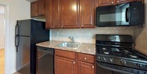 This single-floor unit offers a clean, functional layout with a warm and inviting aesthetic throughout. The living areas are finished with gleaming honey-toned hardwood floors that extend seamlessly through the main rooms, creating a cohesive and bright atmosphere. Large windows dressed with horizontal blinds allow generous natural light to fill the space, while wall-mounted air conditioning units and classic cast-iron radiators ensure year-round comfort. The rooms feature crisp white walls, clean baseboards, and flush-mount ceiling light fixtures that contribute to a simple, uncluttered feel. A closet provides practical storage within the living quarters. The kitchen is outfitted with rich, dark cherry wood cabinetry — both upper and lower — complemented by light stone-finish countertops and a neutral tile backsplash. Black appliances, including a refrigerator, gas range, built-in microwave, and dishwasher, complete the kitchen's functional setup. The bathroom features earthy brown wall tiles surrounding a full soaking tub with sliding glass shower doors, paired with beige floor tiles, a wood-toned vanity, and a wall-mounted medicine cabinet mirror. Overall, the space presents a practical and comfortable living environment with classic finishes throughout.