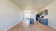 This single-story unit is designed with a functional, open-concept layout that connects the kitchen and living areas seamlessly. The kitchen serves as a central feature, equipped with dark-toned cabinetry, white countertops, and a full suite of black appliances including a refrigerator, gas range, built-in microwave, and dishwasher. A peninsula island with an integrated sink and breakfast bar overhang provides additional workspace and casual seating. Pendant lighting above the island and recessed ceiling lights contribute to a well-lit cooking environment. A window positioned above the sink allows natural light to fill the space.

Light wood-look flooring extends through the kitchen and main living area, complementing the neutral wall tones throughout. The living area is generously sized and includes a ceiling fan with a window that brings in additional natural light. A private entry door is accessible from this area. The bedrooms feature plush carpeting, ceiling fans, and windows with blinds that provide views to the exterior. The floor plan, visible in the 3D scan, reveals a well-organized layout with clearly defined living, kitchen, and bedroom zones, offering a practical and comfortable living environment.