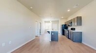 This single-story unit is designed with a functional, open-concept layout that connects the kitchen and living areas seamlessly. The kitchen serves as a central feature, equipped with dark-toned cabinetry, white countertops, and a full suite of black appliances including a refrigerator, gas range, built-in microwave, and dishwasher. A peninsula island with an integrated sink and breakfast bar overhang provides additional workspace and casual seating. Pendant lighting above the island and recessed ceiling lights contribute to a well-lit cooking environment. A window positioned above the sink allows natural light to fill the space.

Light wood-look flooring extends through the kitchen and main living area, complementing the neutral wall tones throughout. The living area is generously sized and includes a ceiling fan with a window that brings in additional natural light. A private entry door is accessible from this area. The bedrooms feature plush carpeting, ceiling fans, and windows with blinds that provide views to the exterior. The floor plan, visible in the 3D scan, reveals a well-organized layout with clearly defined living, kitchen, and bedroom zones, offering a practical and comfortable living environment.