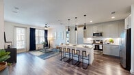 This single-floor residence showcases a thoughtfully designed open-concept layout with wood-style flooring flowing throughout the main living areas. The kitchen serves as a central hub, equipped with stainless steel appliances including a French door refrigerator, range, and microwave. Crisp white shaker cabinetry, quartz countertops, a herringbone tile backsplash, and a generous island with seating for several accommodate both cooking and casual dining. Three sculptural black pendant lights above the island add a contemporary accent. The open living area benefits from abundant natural light through multiple windows dressed with navy curtains, and features a ceiling fan overhead. Bedrooms are carpeted for comfort; the primary suite includes a ceiling fan, dual windows, and a bold navy accent wall. A second bedroom provides flexible use as an additional sleeping space or flex room. The primary bathroom is appointed with a soaking tub framed in marble-style tile, a dual-sink vanity with quartz counters, wood-style flooring, and direct access to a walk-in closet. Throughout, the interior maintains a clean, contemporary aesthetic with neutral tones and modern finishes.