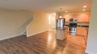 This two-story residence offers a well-designed layout that balances functionality and comfort across both levels. The main floor showcases an open-concept living and kitchen area, creating a spacious and airy atmosphere ideal for both everyday living and entertaining. Rich hardwood flooring flows seamlessly throughout the main level, adding warmth and a sense of continuity to the space.

The kitchen is a standout feature, equipped with stainless steel appliances including a refrigerator, range, and microwave. Light wood cabinetry provides ample storage, while a central island with a dark countertop offers additional workspace and casual seating. Pendant lighting above the island adds a refined touch to the kitchen's overall aesthetic.

Warm, neutral-toned walls complement the natural light that fills the main living area, contributing to a bright and welcoming ambiance. A staircase with clean white railings leads to the second floor, hinting at additional living or sleeping quarters above. A utility or storage area is also visible off the main corridor, adding to the home's practical appeal. Overall, the space presents a clean, modern design with thoughtful details throughout.