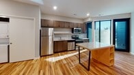 This well-appointed single-floor residence offers a thoughtfully designed layout with modern finishes throughout. The kitchen serves as a central highlight, featuring rich dark wood cabinetry, a crisp white tile backsplash, stone countertops, and a full suite of stainless steel appliances including a refrigerator, gas range, microwave, and dishwasher. A kitchen island with a stone surface provides additional prep and dining space. Large windows flood the kitchen and adjacent areas with generous natural light, enhancing the warm tones of the hardwood flooring that extends throughout the living spaces. The open layout offers a versatile living area complete with recessed built-in shelving, ideal for display or storage. A dedicated in-unit laundry area with a stacked washer and dryer adds everyday convenience. The bathroom is finished with a wood vanity, granite-style countertop, an undermount sink, a large mirror, and open wooden shelving for organized storage. Recessed lighting throughout the space provides a clean, modern ambiance, while crisp white walls contrast beautifully with the warm wood tones to create a bright and welcoming environment.
