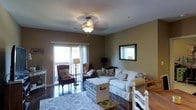 This single-floor space presents a warm and inviting atmosphere, characterized by neutral earth-toned walls that create a cozy, relaxed ambiance throughout. The living area is thoughtfully arranged with a white slipcovered sofa accented by decorative pillows, complemented by classic wingback chairs in rich upholstery, offering ample seating for relaxation. A distinctive white vintage trunk serves as a functional coffee table centerpiece, adding a charming, eclectic touch to the room. Dark hardwood floors run throughout, providing an elegant contrast to the lighter furnishings and a patterned area rug that anchors the seating arrangement. Natural light fills the space generously through large sliding glass doors that open to an outdoor balcony, offering views of the surrounding landscape. A framed mirror mounted on the wall enhances the sense of space and reflects additional light into the room. The ceiling features a classic light fixture with a ceiling fan, ensuring both comfort and illumination. A secondary window on an adjacent wall further brightens the interior. The layout seamlessly blends living and sleeping areas, making thoughtful use of the available space while maintaining a cohesive, comfortable aesthetic throughout.