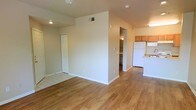 This single-story space offers an open, functional layout with hardwood-style flooring extending throughout the main living areas. The open-concept design seamlessly connects the living area to the kitchen, creating a cohesive and practical flow. The kitchen is equipped with wood-toned cabinetry, a white refrigerator, and light countertops, and is positioned at the far end of the space for easy accessibility. A central dividing wall adds subtle definition between zones without closing off the layout. Recessed and overhead lighting fixtures are installed throughout, ensuring consistent illumination across the interior. Neutral, light-toned walls contribute to a bright and airy atmosphere. A doorway visible in the background leads to an additional room or hallway, suggesting further utility spaces beyond the main area. A closet with white bifold doors is situated along one wall, providing built-in storage. The transition between tile and hardwood-style flooring near the closet area subtly delineates separate functional zones. Overall, the space presents a clean, straightforward interior well-suited for everyday living.