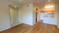 This single-story space offers an open, functional layout with hardwood-style flooring extending throughout the main living areas. The open-concept design seamlessly connects the living area to the kitchen, creating a cohesive and practical flow. The kitchen is equipped with wood-toned cabinetry, a white refrigerator, and light countertops, and is positioned at the far end of the space for easy accessibility. A central dividing wall adds subtle definition between zones without closing off the layout. Recessed and overhead lighting fixtures are installed throughout, ensuring consistent illumination across the interior. Neutral, light-toned walls contribute to a bright and airy atmosphere. A doorway visible in the background leads to an additional room or hallway, suggesting further utility spaces beyond the main area. A closet with white bifold doors is situated along one wall, providing built-in storage. The transition between tile and hardwood-style flooring near the closet area subtly delineates separate functional zones. Overall, the space presents a clean, straightforward interior well-suited for everyday living.