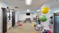 Located at 2500 Edwards Dr in Fort Myers, Florida, this single-floor rehabilitation and physical therapy facility is thoughtfully designed to support a wide range of therapeutic and exercise-based programs. The space features warm hardwood flooring throughout, white walls, and drop-tile ceilings with recessed fluorescent lighting that create a clean, clinical yet welcoming environment.

The centerpiece of the facility is a generously sized open exercise area that provides ample room for movement and group activities. This main space is equipped with wall-mounted suspension training bars — including TRP straps and a speed bag — as well as a rack of free weights and parallel bars for balance and gait training. Storage benches along the walls neatly organize an assortment of stability balls in various sizes and colors, resistance bands, exercise mats, and other therapeutic tools.

An adjoining area, separated by a partial wall, extends the usable floor space and houses additional rehabilitation equipment including foam rollers, exercise hoops, and modular mats, making it well suited for stretching routines or small-group sessions.

Smaller individual rooms branch off from the main exercise space, offering more private settings for one-on-one consultations or focused therapeutic work. These rooms feature the same consistent hardwood floors and neutral tones, maintaining a cohesive and professional atmosphere throughout the entire facility.