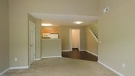 This two-story residence offers a practical and comfortable layout designed for everyday living. The main floor centers around an open living area finished with plush wall-to-wall carpeting, creating a warm and inviting atmosphere. Neutral sage-green walls carry throughout the space, lending a cohesive and calming aesthetic. The kitchen is positioned adjacent to the living area and is defined by a raised bar-height peninsula counter, providing a natural visual separation between the two spaces. Warm wood-toned cabinetry lines the kitchen walls above, and appliances including a microwave and refrigerator are neatly integrated. Dark hardwood flooring in the kitchen and connecting hallway offers a striking contrast to the carpeted living space. A doorway off the main area leads to an additional room, adding versatility to the floor plan. The staircase, positioned along one wall, features clean white railings and leads to the second floor, where additional living and sleeping spaces are found. Flush-mount ceiling lighting provides consistent illumination throughout. White trim and baseboards frame each room cleanly, adding a polished finish to the overall design.