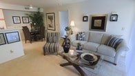 This single-floor residence offers an open, thoughtfully designed layout that connects the living and dining areas in a cohesive flow. The living room serves as the centerpiece, featuring plush, neutral-toned seating including a generously sized sofa and a striped accent chair that add visual interest without overwhelming the understated palette. A glass-topped coffee table with a sculptural wooden base anchors the seating arrangement, contributing a refined, contemporary touch.

The walls are painted in soft, warm neutrals, creating a calm and inviting atmosphere throughout. Framed artwork adorns the walls, complementing the carefully curated décor that includes decorative vases, potted orchids, and lush indoor plants that introduce natural elements into the space. Warm ambient lighting from table lamps enhances the cozy feel of the room.

Adjacent to the living area, the dining space accommodates a dark rattan table and chairs, grounded by the same light-toned carpeting that runs throughout. The consistent flooring and color scheme unify the open-plan layout, giving the space a sense of cohesion and generous scale.