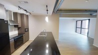 This contemporary single-floor space showcases a thoughtfully designed open-concept layout that blends modern aesthetics with industrial character. The kitchen serves as a central highlight, anchored by a generous dark granite island with a polished finish and an integrated stainless steel sink equipped with a high-arc faucet. Pendant lighting hangs above the island, adding a refined focal point to the space. Dark wood cabinetry lines the kitchen wall, complemented by built-in appliances including a microwave and oven, offering both style and functionality. Track lighting runs along the ceiling, providing flexible and even illumination throughout the kitchen area. A standout industrial feature is the exposed ductwork that runs prominently along the ceiling, lending the space an urban loft character. Light wood-toned flooring extends throughout, creating a warm contrast to the darker finishes. The kitchen opens directly into a bright, airy living area, where large windows allow natural light to fill the space, enhancing its open and inviting atmosphere. The overall design strikes a balance between modern sophistication and industrial edge, making it a versatile and visually compelling living environment.