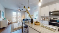 This single-floor residence offers a thoughtfully designed open-concept layout that seamlessly connects the kitchen, dining, and living areas. The kitchen is a standout feature, showcasing white shaker-style cabinetry, granite countertops, a large peninsula with bar seating illuminated by pendant lighting, and stainless steel appliances including a refrigerator, range, and microwave. Light wood-style flooring runs throughout the main living areas, enhancing the bright and airy atmosphere. The living room provides a comfortable gathering space with a ceiling fan, access to a private outdoor area through French doors, and ample natural light from large windows. The space includes multiple bedrooms, each carpeted and filled with natural light courtesy of large windows with blinds and curtains. The primary bedroom features a spacious walk-in closet and an en-suite bathroom with a tub-shower combination. A second full bathroom features a white vanity with granite countertop, a large mirror, and hardwood-style flooring. The overall aesthetic is clean and contemporary, with a cohesive palette of whites, grays, and soft blues that creates an inviting and functional living environment throughout.