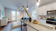 This single-floor residence offers a thoughtfully designed open-concept layout that seamlessly connects the kitchen, dining, and living areas. The kitchen is a standout feature, showcasing white shaker-style cabinetry, granite countertops, a large peninsula with bar seating illuminated by pendant lighting, and stainless steel appliances including a refrigerator, range, and microwave. Light wood-style flooring runs throughout the main living areas, enhancing the bright and airy atmosphere. The living room provides a comfortable gathering space with a ceiling fan, access to a private outdoor area through French doors, and ample natural light from large windows. The space includes multiple bedrooms, each carpeted and filled with natural light courtesy of large windows with blinds and curtains. The primary bedroom features a spacious walk-in closet and an en-suite bathroom with a tub-shower combination. A second full bathroom features a white vanity with granite countertop, a large mirror, and hardwood-style flooring. The overall aesthetic is clean and contemporary, with a cohesive palette of whites, grays, and soft blues that creates an inviting and functional living environment throughout.