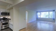 This single-floor space is characterized by its clean, minimalist design and functional layout. The main living area features light-toned hardwood flooring that extends throughout, contributing to a bright and open atmosphere. Neutral white walls provide a versatile backdrop, while a large window along the far wall invites abundant natural light and offers views of surrounding greenery, enhancing the sense of openness. A baseboard heating unit runs beneath the window, ensuring practical comfort. A doorway visible within the space leads to an adjoining room, suggesting a straightforward and accessible floor plan. The low-profile ceiling and uncluttered finishes give the space a modest yet tidy character. Overall, the space presents a simple, well-lit environment with quality flooring and adequate natural light, making it a practical and adaptable living area.