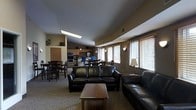 This single-floor community space is thoughtfully designed to accommodate a variety of activities within an open, flowing layout. The lounge area anchors the space with dark leather sofas arranged around a wooden coffee table, creating a welcoming setting for relaxation and conversation. Natural light filters in through multiple windows fitted with horizontal blinds, brightening the neutral-toned walls and carpeted flooring throughout.

Adjacent to the lounge, a fully equipped kitchen offers stainless steel appliances, including a refrigerator, oven, and microwave, set against warm wood-toned cabinetry. A dining area with multiple tables and chairs sits nearby, providing ample seating for group meals or activities. Wall-mounted light sconces and a floor lamp contribute to a warm, ambient atmosphere during evening hours.

The open-concept design ensures seamless transitions between the lounge, dining, and kitchen zones, making the space highly functional for both casual use and organized gatherings. Recessed ceiling details and varied ceiling heights add architectural interest, while the consistent use of warm earth tones throughout creates a cohesive and inviting environment.