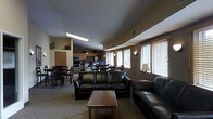 This single-floor community space is thoughtfully designed to accommodate a variety of activities within an open, flowing layout. The lounge area anchors the space with dark leather sofas arranged around a wooden coffee table, creating a welcoming setting for relaxation and conversation. Natural light filters in through multiple windows fitted with horizontal blinds, brightening the neutral-toned walls and carpeted flooring throughout.

Adjacent to the lounge, a fully equipped kitchen offers stainless steel appliances, including a refrigerator, oven, and microwave, set against warm wood-toned cabinetry. A dining area with multiple tables and chairs sits nearby, providing ample seating for group meals or activities. Wall-mounted light sconces and a floor lamp contribute to a warm, ambient atmosphere during evening hours.

The open-concept design ensures seamless transitions between the lounge, dining, and kitchen zones, making the space highly functional for both casual use and organized gatherings. Recessed ceiling details and varied ceiling heights add architectural interest, while the consistent use of warm earth tones throughout creates a cohesive and inviting environment.