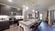This thoughtfully designed single-floor residence offers a seamless blend of modern style and functional living. The open-concept kitchen is a standout feature, showcasing dark espresso cabinetry that extends to the ceiling, subway tile backsplash with under-cabinet lighting, stainless steel appliances, and granite countertops. A large central island with an integrated sink and bar seating serves as both a prep space and a casual dining hub, illuminated by elegant pendant lights overhead. Wood-look flooring flows throughout the main living areas, anchoring the cohesive aesthetic.

The living room is a comfortable and inviting space, filled with natural light from large windows dressed with decorative curtains. A glass-paneled door provides additional brightness and access to the outdoors. The bedroom continues the warm, residential feel with plush carpeting, a ceiling fan, and generously sized windows that frame exterior views. The en-suite bathroom is well-appointed with granite countertops, dark cabinetry, wood-look tile flooring, and direct access to a spacious walk-in closet with built-in shelving and hanging storage. Together, the rooms present a well-balanced, contemporary living environment with quality finishes throughout.