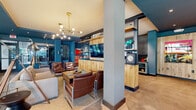 This thoughtfully designed single-floor communal lounge delivers a dynamic blend of industrial and contemporary aesthetics. Bold teal-painted walls and ceilings create a striking visual identity throughout, contrasted by warm natural wood paneling and open shelving that anchor the central media and lounge zone. Polished concrete floors extend seamlessly across the entire space, lending an urban, loft-like quality underfoot.

The main lounge area is furnished with a generous sectional sofa, leather armchairs with chrome frames, and a wooden coffee table, all arranged atop a patterned area rug. Overhead, a multi-arm brass Sputnik chandelier adds a retro-modern focal point, while industrial-style floor lamps on tripod bases contribute to the eclectic, curated atmosphere. Open shelving behind the seating area displays decorative objects alongside a wall-mounted flat-screen television.

A dedicated co-working and bar-height seating area lines the perimeter, positioned directly in front of expansive floor-to-ceiling windows that flood the space with natural light and offer clear views of the surrounding streetscape. Blue counter stools line a long wall-mounted counter, making it an ideal spot for individual work or casual gatherings. Additional seating nooks with upholstered wingback chairs and natural wood stump side tables provide quieter corners for relaxed conversation. The open layout and abundant glazing give the entire space an airy, connected, and welcoming feel.