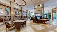 Located in King of Prussia, PA, this impressive clubhouse spans a single, generously proportioned floor and offers a wide array of thoughtfully designed spaces for gathering and recreation. The grand central lounge serves as the heart of the facility, anchored by a dramatic floor-to-ceiling glass-paneled fireplace and furnished with richly upholstered seating arranged on patterned area rugs over marble-tiled flooring. Built-in bookshelves line the walls, complemented by curated artwork and warm, layered lighting from statement chandeliers and pendant fixtures. Adjacent to the lounge is a cozy library retreat, complete with a traditional fireplace, floor-to-ceiling bookshelves, and plush seating that creates an inviting, intimate atmosphere. A dedicated game room provides entertainment with ping pong tables, a billiards area, a wall-mounted TV, and bold navy cabinetry beneath a warm wood-paneled ceiling. A stylish café-style kitchen features a striking marble island, herringbone-patterned hardwood flooring, and sophisticated brass-accented shelving. A bright dining lounge with coffered wood ceilings and large windows opens directly to a landscaped outdoor patio with seating. The elevator lobby showcases herringbone hardwood floors and elegant wood-beam ceilings, tying together the refined architectural character throughout this exceptional clubhouse.