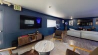 This single-floor space makes a bold statement with its deep navy blue walls, white ceilings, and wood-look flooring that run throughout, creating a cohesive and moody aesthetic. The main living and entertainment area is generously sized, anchored by a large wall-mounted flat-screen TV, a rustic wood media console with a record player, and an assortment of eclectic seating including mid-century style armchairs arranged atop cowhide and patterned area rugs. A classic popcorn machine adds a fun, lounge-like touch to this entertainment zone. An iconic blue neon sign reading "This must be the place" serves as a striking focal point in the adjoining lounge and dining area, which features a solid wood dining table and ample seating. Natural light filters in through multiple windows fitted with white blinds throughout the space. A dedicated mirrored room offers floor-to-ceiling mirror panels, wood-look flooring, a white reception counter with a stone-finish surface, and track lighting overhead — ideal as a studio or creative workspace. The kitchen is clean and functional, featuring white shaker-style cabinetry, stone countertops, a black gas range, and a glass-panel entry door that floods the room with natural light.