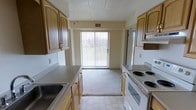 This single-floor space features a galley-style kitchen that serves as the central hub of the layout. The kitchen is equipped with warm-toned wooden cabinetry lining both sides of the corridor, providing generous storage throughout. Laminate countertops run along each wall, complemented by a built-in sink on one side and a white electric coil-burner range with an overhead exhaust hood on the other. The tile flooring in the kitchen transitions smoothly into carpeted living space beyond.

A standout feature of this space is the sliding glass door at the end of the kitchen corridor, fitted with vertical blinds that allow for adjustable natural light. This door opens to an outdoor area with visible greenery, creating a pleasant visual connection to the outside. The natural light that flows in through this door brightens the kitchen and adjacent living area considerably.

The walls are finished in a neutral, warm beige tone, contributing to an inviting atmosphere. The textured ceiling adds a classic touch. A ceiling vent ensures climate control throughout. Overall, the space offers a straightforward and practical design suited for comfortable everyday living.