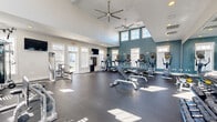 Located at Coventry Commons in Concord, NC, this dedicated fitness center offers a generously sized, single-floor exercise space designed to support a full range of workout routines. The room is defined by its striking vaulted ceilings, which lend an open, airy feel, and are complemented by dual large ceiling fans that promote air circulation throughout the space. A distinctive teal accent wall anchors the room visually, contrasting cleanly against crisp white walls and ceilings.

Natural light pours in through multiple windows, including a set of elevated clerestory windows along the accent wall, creating a bright and energizing environment. The durable dark rubber flooring provides both safety and comfort underfoot.

The fitness center is thoroughly equipped for cardiovascular and strength training. Cardio equipment includes multiple treadmills, elliptical trainers, and stationary bikes, several of which feature individual screens. Strength training options include a cable machine, multi-station weight apparatus, adjustable benches, and a full rack of dumbbells and kettlebells. Wall-mounted flat-screen televisions are positioned throughout for entertainment. A large wall mirror expands the sense of space while allowing users to monitor their form. The overall layout is thoughtfully organized to maximize usability and flow.