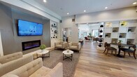This well-appointed single-floor community clubhouse offers a thoughtfully designed layout with distinct areas for relaxation, entertainment, and socializing. The central lounge area features a wall-mounted flat-screen TV above a sleek electric fireplace, flanked by built-in glass shelving displaying decorative accents. Comfortable upholstered seating is arranged around marble-topped coffee tables atop a patterned area rug, all set on warm hardwood flooring that flows throughout the space. Adjacent to the lounge, a dedicated media and screening room provides a large projection screen framed by a custom built-in surround, overhead projector, stylish wall sconces, and a generous sectional sofa paired with bistro-height seating for a versatile viewing experience. A bright dining area benefits from expansive gridded windows that invite abundant natural light and views of the surrounding greenery, with a substantial wood dining table and upholstered chairs beneath soaring ceilings. A separate sitting room with floor-to-ceiling windows and a neutral palette creates a serene retreat for conversation. The kitchen features granite countertops, subway tile backsplash, stainless steel appliances, and a dedicated coffee bar station, completing this comprehensive and elegantly appointed communal facility.