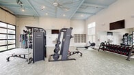 Located at 420 Angler Drive in Wilmington, NC, this well-appointed fitness center offers a generous, open exercise space designed to support a full range of workout routines. The room is defined by its striking vaulted ceiling with exposed light-blue painted beams, recessed lighting, and a large ceiling fan that keeps the space comfortable during intense sessions. Tall windows and full-panel glass garage doors flood the interior with natural light and provide open views of the surrounding community, creating an inviting and airy atmosphere. The flooring consists of durable, textured carpet suited for fitness use throughout the entire space. Equipment includes a cable machine system with an integrated Instinct strength trainer, a weight bench with free weights, a rowing machine, dumbbells on a dedicated rack, kettlebells, medicine balls, resistance bands, elliptical trainers, stationary bikes, and side-by-side treadmills. Wall-mounted flat-screen TVs are positioned for easy viewing from multiple workout stations. A built-in water fountain station with decorative tile accent wall, open cubby storage shelving, and access doors to additional rooms add practical convenience. The overall design balances functionality with a bright, modern aesthetic.
