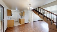 This two-story space offers a functional and straightforward layout across two well-defined levels. The main floor features warm hardwood flooring that extends through the open living area, anchored by a classic chandelier that adds a touch of character overhead. A staircase with dark wood treads and black metal balusters serves as a prominent architectural element, connecting the two floors with a clean, open design. The galley-style kitchen is efficiently arranged with warm oak cabinetry lining both sides, light countertops, tile flooring, and white appliances including a range and refrigerator, alongside a window that brings in natural light. A door visible from the main living area provides additional access. The upper level hosts carpeted bedrooms, each featuring windows that frame views of surrounding greenery and neighboring structures, filling the rooms with natural light and a sense of openness. The bathrooms are finished with neutral, light-toned tile surrounding a full soaking tub, and a vanity with a large mirror and light countertop provides practical utility. Throughout the space, a neutral color palette of whites and warm wood tones creates a clean, cohesive atmosphere that is both functional and inviting.