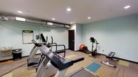 This fitness center offers a practical and functional workout environment designed to accommodate a variety of exercise routines. The space is finished with light hardwood-style flooring accented by colorful exercise mat areas in blue and yellow, providing designated zones for stretching and floor exercises. The walls are painted in a cool, muted teal tone that contributes to a calm and focused atmosphere. Recessed lighting illuminates the room evenly, complemented by exposed ventilation ductwork along the ceiling that adds an industrial character to the space. The cardio equipment selection includes treadmills and elliptical machines positioned toward one side of the room, while a stationary upright bike occupies the opposite corner. An exercise mat and stability ball are also available, supporting core training and flexibility work. A wall-mounted informational board near the entrance provides guidance for users. A dark-framed door leads out of the space, keeping the layout clean and unobstructed. The overall design prioritizes functionality and ease of movement, making it a well-rounded facility suitable for a range of fitness levels and workout styles.