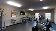 Located at 300 Northwoods Rd in Middletown, NY, this fitness center offers a well-equipped, single-floor exercise space designed to support a well-rounded workout routine. The room features a dark, durable flooring surface suited for active use, with neutral-toned walls that create a focused and functional atmosphere. Natural light filters in through a window dressed with a patterned valance, complementing the overhead fluorescent lighting that keeps the space bright and welcoming. A wall-mounted flat-screen television is positioned for easy viewing during workouts, accompanied by a digital clock display for time management. A full-length wall mirror along one side of the room enhances the sense of openness and allows users to monitor their form. The free weight section includes a well-organized multi-tier dumbbell rack stocked with a range of weights, along with a dedicated bench area. The cardio section features an elliptical trainer, a stationary bike, and a treadmill, providing a variety of aerobic options. A vintage-style spa and cycling poster adds a touch of character to the walls. The space is thoughtfully arranged to maximize usability while maintaining clear, open floor space for stretching or floor exercises.