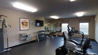 Located at 300 Northwoods Rd in Middletown, NY, this fitness center offers a well-equipped, single-floor exercise space designed to support a well-rounded workout routine. The room features a dark, durable flooring surface suited for active use, with neutral-toned walls that create a focused and functional atmosphere. Natural light filters in through a window dressed with a patterned valance, complementing the overhead fluorescent lighting that keeps the space bright and welcoming. A wall-mounted flat-screen television is positioned for easy viewing during workouts, accompanied by a digital clock display for time management. A full-length wall mirror along one side of the room enhances the sense of openness and allows users to monitor their form. The free weight section includes a well-organized multi-tier dumbbell rack stocked with a range of weights, along with a dedicated bench area. The cardio section features an elliptical trainer, a stationary bike, and a treadmill, providing a variety of aerobic options. A vintage-style spa and cycling poster adds a touch of character to the walls. The space is thoughtfully arranged to maximize usability while maintaining clear, open floor space for stretching or floor exercises.