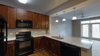 This single-floor space centers around a thoughtfully designed kitchen that combines functionality with classic style. The kitchen is outfitted with rich, dark cherry wood cabinetry featuring brushed nickel hardware, offering generous storage along both upper and lower cabinet runs. Light-toned granite countertops provide ample workspace and contrast warmly against the dark cabinetry. Black appliances, including an electric range with oven, built-in microwave, and dishwasher, lend a cohesive, modern look throughout the cooking area. A double stainless steel sink is positioned at the peninsula counter, which opens directly toward the adjacent living area, creating an open, connected layout ideal for entertaining. Three pendant lights with white dome shades hang above the peninsula, providing focused task lighting while enhancing the visual transition between the kitchen and living space. The hard wood-style flooring in the kitchen grounds the space with warmth and durability. The adjoining room, visible through the open pass-through, features neutral gray walls and carpeted flooring, creating a comfortable and cohesive flow throughout the home's single level.