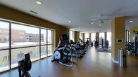 This fitness center offers a spacious, single-floor layout designed to accommodate a comprehensive workout experience. The room features warm, earthy-toned walls paired with hardwood-style flooring, creating an inviting yet functional atmosphere. Large floor-to-ceiling windows line one side of the space, flooding the interior with abundant natural light while providing views of the surrounding urban landscape. Additional windows at the far end of the room further enhance the bright, airy feel.

The cardio section is well-stocked with a row of elliptical trainers, treadmills, and stationary bikes, all aligned to take advantage of the natural light and views. Toward the rear of the space, strength training equipment and cable machines are available for resistance-based workouts. Ceiling fans are installed throughout, contributing to comfortable air circulation during exercise sessions.

Recessed lighting complements the natural light, ensuring the space remains well-lit at all times. The open layout provides ample room to move between equipment, making the fitness center feel accessible and easy to navigate for users of all fitness levels.