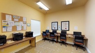 This single-floor business center offers a practical and well-equipped workspace designed to meet a variety of productivity needs. The room features warm yellow walls that create an inviting atmosphere, complemented by a patterned carpet that adds texture and visual interest underfoot. Overhead, recessed fluorescent lighting ensures the space is consistently and evenly illuminated throughout.

Along one wall, a built-in wooden desk runs the length of the room, providing ample surface space for multiple computer workstations. Each station is outfitted with a monitor and ergonomic office chair upholstered in black and orange, offering comfortable seating for extended use. A dedicated printer and copier station is positioned near a window fitted with white horizontal blinds, allowing for natural light control as needed.

The walls are tastefully decorated with framed artwork, adding a touch of warmth to the functional environment. A corkboard near the entrance displays informational notices and announcements for easy reference. A security camera mounted near the ceiling ensures the safety of the space. Overall, the business center balances functionality with a welcoming design, making it a versatile resource for visitors or residents.