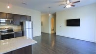 This single-floor space showcases an open-concept design that seamlessly connects the kitchen and living areas, creating a fluid and functional layout. The dark hardwood flooring runs throughout, providing a rich contrast against the light-toned walls and adding a sleek, contemporary feel to the interior.

The kitchen is equipped with dark cabinetry, a stainless steel refrigerator, and a granite-topped island that doubles as a casual dining or prep surface. A pendant light hangs above, adding a focused decorative touch to the space. The kitchen transitions naturally into the main living area, which features a ceiling fan with integrated lighting and a wall-mounted flat-screen television, making it a practical and comfortable gathering space.

A hallway visible from the main area leads to a bathroom, offering clear spatial organization throughout the unit. The neutral wall palette keeps the atmosphere clean and versatile, allowing for a variety of interior styling options. White baseboards provide a crisp finish throughout. Overall, the space presents a modern, low-maintenance aesthetic suitable for everyday living.