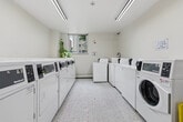 Located at 80 Cosburn Ave in Toronto's East End, this shared laundry facility offers a clean, well-organized space designed for practical, everyday use. The room is finished with classic white mosaic tile flooring and light-toned walls that contribute to a bright, sanitary atmosphere. Natural light filters in through a window overlooking the surrounding neighbourhood, complemented by overhead fluorescent lighting that ensures the space is well-lit at all hours. Multiple top-loading and front-loading washers and dryers line the walls, providing ample capacity for building residents. A dedicated utility sink with a faucet offers added convenience for hand-washing or pre-treating garments. The room also features a pet-friendly machine, catering to residents with animals. A fire extinguisher and security camera are present, reflecting a commitment to safety. Additional amenities include folding surfaces and clearly posted usage guidelines to help keep the space tidy and orderly. Easy access through a central doorway connects the laundry room to the rest of the building, making it a highly functional and accessible shared amenity for all residents.