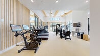 This fitness center is a thoughtfully designed, single-floor facility that blends contemporary aesthetics with comprehensive workout functionality. The space is defined by soaring ceilings with exposed wood beam detailing and ceiling fans with gold accents, contributing to an open, airy atmosphere throughout. Vertical wood slat wall panels add warmth and texture, complementing the clean white walls and light-toned flooring underfoot.

Large black-framed floor-to-ceiling windows and glass doors line the perimeter, flooding the interior with abundant natural light and offering views of the surrounding greenery and outdoor areas. The bright, open layout is divided into distinct functional zones. The cardio section is well-appointed with treadmills, stationary bikes with integrated display screens, and stair climbers. The strength training area features a comprehensive selection of cable machines, weight machines, and a full rack of dumbbells.

A dedicated free weights and stretching zone provides ample room for flexibility and functional training, with stability balls and mats stored on wall-mounted racks. Amenity details include a wall-mounted television, a water fountain station, and a compact refrigeration unit. The overall ambiance is clean, modern, and inviting, making this fitness center a well-rounded facility suited for a wide range of training needs.