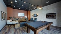 This single-floor entertainment lounge is thoughtfully designed to offer a dynamic and social atmosphere. The space centers around a full-size pool table with a sleek dark felt surface, positioned beneath a modern linear pendant light that anchors the room visually. The ceiling features a bold dark grid framework with recessed track lighting, adding an industrial yet polished aesthetic throughout.

The lounge is divided into distinct activity zones. A comfortable seating area includes plush gray armchairs and a sofa arranged around a low coffee table, accompanied by a wall-mounted flat-screen TV — ideal for relaxed viewing or conversation. An additional TV is mounted on the striking textured stone accent wall, which serves as a dramatic focal point and contrasts beautifully with the warm terracotta-toned painted walls on the opposite side.

A kitchenette area along one wall is equipped with a microwave, beverage refrigerator, and cabinetry, providing convenient amenities for entertaining. The flooring features a multi-toned patterned carpet that adds warmth and visual interest underfoot. Together, the varied textures, layered lighting, and well-appointed furnishings create a vibrant yet comfortable gathering space suited for recreation and socializing.